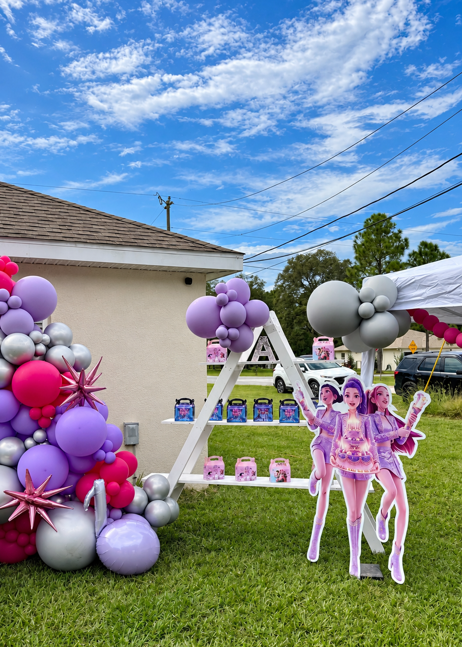 Custom K-pop birthday backdrop with pink, purple and silver balloon décor, character cutouts and white pedestals.