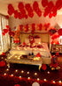 Romantic bedroom decorated with red heart balloons, rose petals, LED lights, teddy bear, champagne, and chocolates for love package celebration