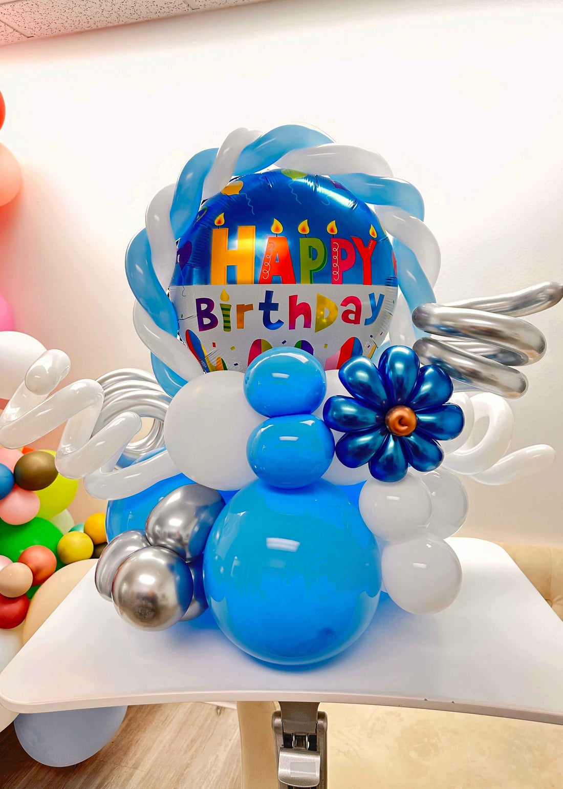 Colorful Birthday Twirl Balloon bouquet with blue, white, and silver balloons and a "Happy Birthday" foil balloon centerpiece