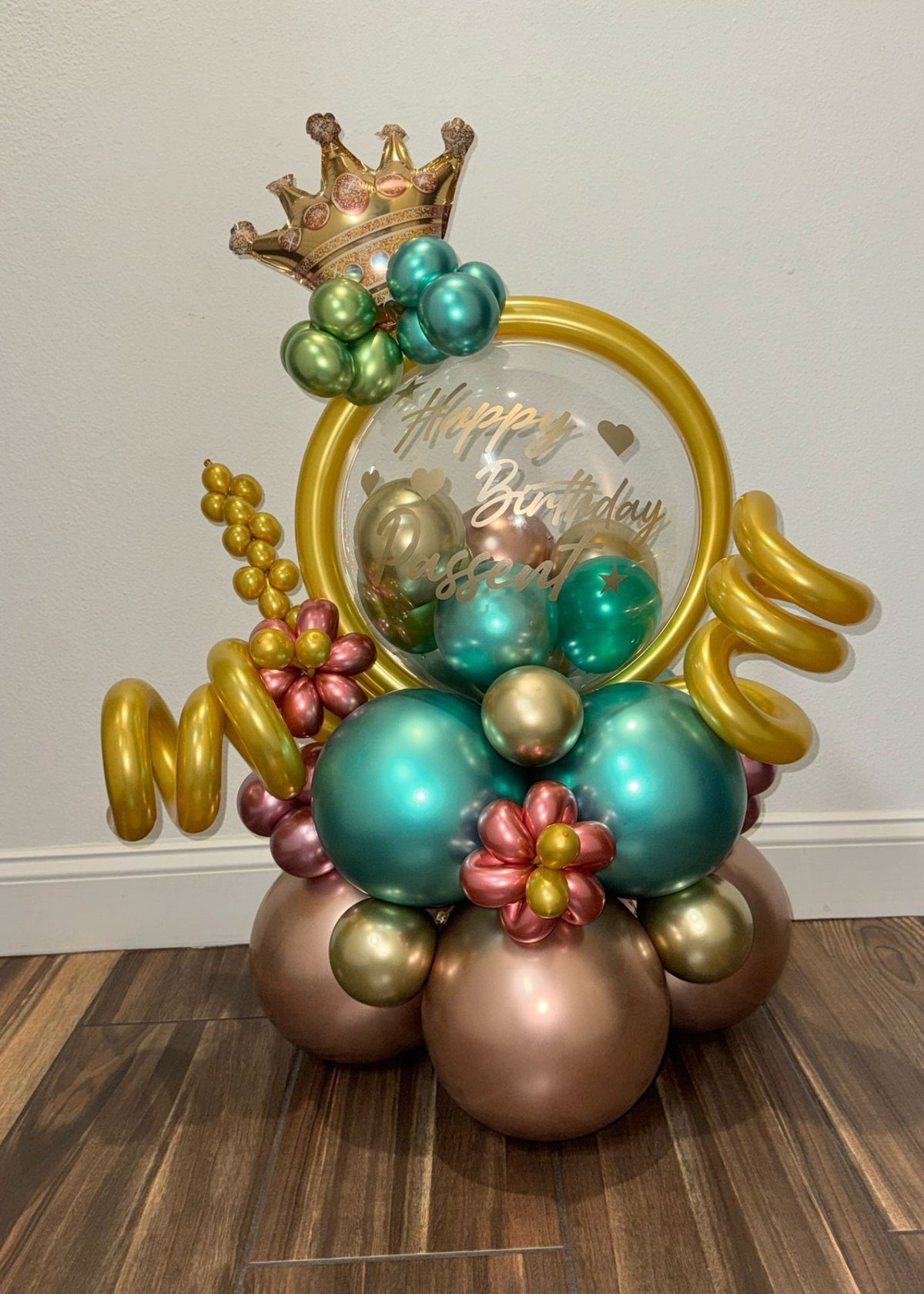 Regal birthday balloon bouquet with gold crown topper, metallic teal, gold, rose pink, bronze balloons, and curled golden ribbons